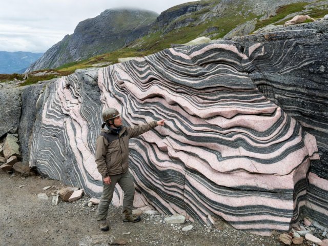 Geology educator pointing at tightly folded pink and dark grey migmatitic gneiss in a Norwegian mountain roadcut