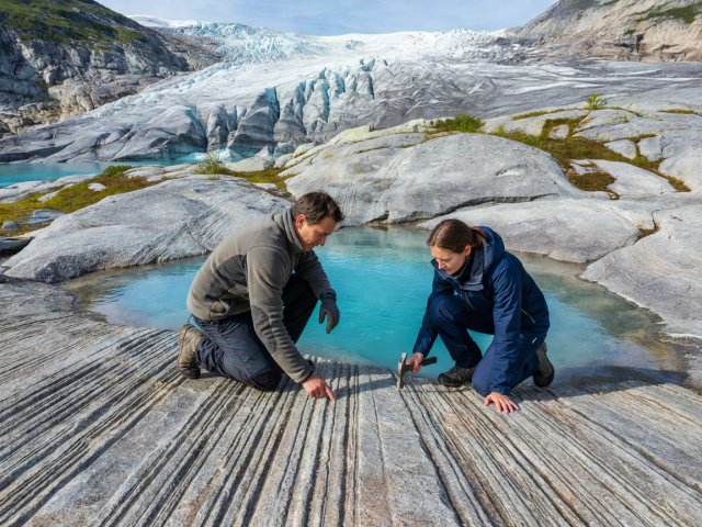 Two hikers kneeling on smoothed gneiss bedrock near Jostedalsbreen examining glacial striations and turquoise meltwater